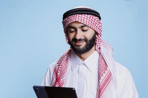 Smiling man wearing arabic clothes using digital tablet and scrolling social media. Cheerful muslim person dressed in ghutra headscarf and thobe tapping on portable device screen photo