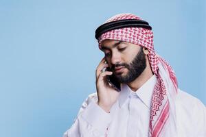 Man wearing arab thobe and headscarf speaking on mobile phone with neutral face expression. Muslim person in islamic headdress speaking on smartphone while posing in studio photo