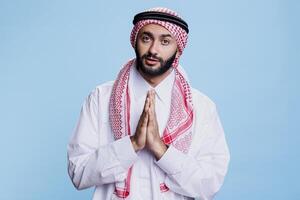 Man wearing traditional muslim thobe and ghutra clothes praying studio portrait. Arab person standing with folded hands showing symbol of faith and spirituality while looking at camera photo