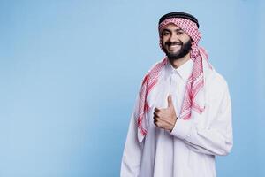 Muslim man dressed in traditional clothes showing thumb up while standing with cheerful expression portrait. Smiling islamic person confirming approval and looking at camera photo