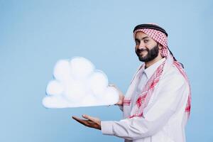 Cheerful man dressed in muslim traditional clothes showing white cloud shape and looking at camera with smiling expression. Carefree arab holding cardboard in studio portrait on blue background photo