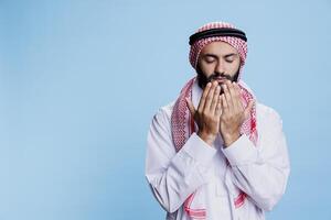 musulmán hombre vistiendo tradicional ropa en pie con abierto palmas mientras Orando. religioso árabe oración en thobe y Pañuelo posando con cerrado ojos mientras preguntando Dios para bendición foto