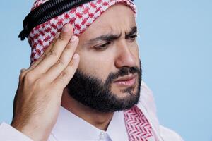 Muslim man rubbing temple and suffering from migraine symptom with tired expression. Arab person wearing thobe and ghutra traditional clothes having headache and feeling pain photo