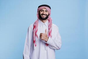Arab man wearing islamic traditional clothes posing with thumb up signifying approval and agreement portrait. Smiling muslim person in thobe and ghutra showing like gesture photo