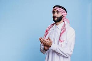 Serious arab man dressed in traditional clothes applauding and showing support portrait. Muslim person clapping hands and looking at camera with confident facial expression photo