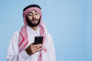 Muslim man wearing traditional clothes texting on smartphone while posing in studio. Arab person dressed in thobe and ghutra sending online message on mobile phone on blue background photo