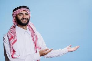 Smiling muslim man showcasing product by pointing to side with arms and looking at camera with cheerful expression. Arab dressed in islamic thobe and headscarf, pointing at left direction photo