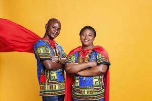 Smiling black couple dressed in superhero red cloaks posing with folded hands and looking at camera. Cheerful superman and superwoman pair wearing capes showing power and strength portrait photo