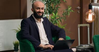 Middle eastern man typing on laptop while relaxing in lobby area and writing speech for global company event. Professional guest travelling on business trip, hotel reception. photo