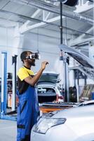 Repairman in auto repair shop using virtual reality to visualize car fuel tank in order to fix it. BIPOC adept garage worker wearing futuristic vr headset while working on busted vehicle photo