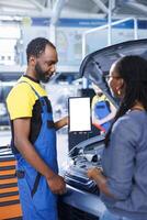 Smiling engineer in car service uses mockup tablet to order new compressor belt for damaged vehicle. Employee showing client needed components replacement on isolated screen device photo