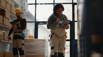 Relaxed african american warehouse picker listening music while checking labels on cardboard boxes. Smiling worker wearing heaphones and bopping head while walking through storage area aisles photo