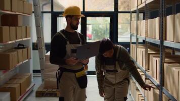 Employee and helpful colleague inputing updated products info on online shopping website, walking through warehouse storage center rows checking labels on cardboard boxes sitting on shelves photo