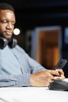 African american guy texting messages on phone app, working from home at his workstation. Male freelancer student checking websites and listening to online training program, e learning. photo