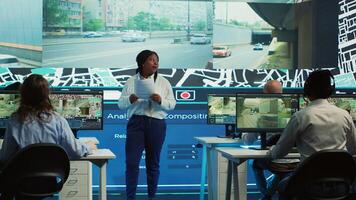 African american manager provides information for delivery parcels to her diverse team, people monitoring traffic on routes. Order tracking in control room, satellite maps with coordinates. Camera A. photo