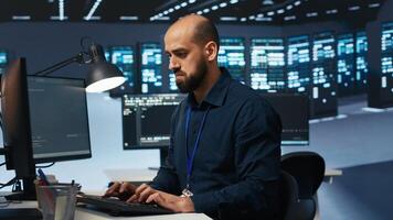 Engineer coding, working in network security data center facility hosting supercomputers, ensuring optimal performance. IT expert using desktop PC to monitor energy consumption across server cabinets photo