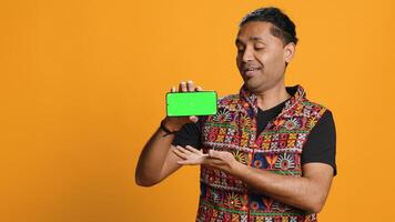 Content creator presenting green screen mobile phone, isolated over studio background. Indian person reviewing copy space mockup smartphone device for internet channel, camera A photo