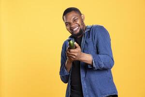 African american funny man pretending to use a cucumber as a shotgun in studio, playing around and having fun with eco friendly vegetables. Model acting goofy, zero waste concept. photo