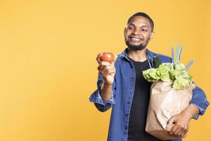 joven persona sonriente y posando con un maduro Fresco tomate, admirativo eco simpático Produce desde local tienda de comestibles almacenar. modelo participación un papel bolso lleno de abultar productos, éticamente de origen verduras. foto