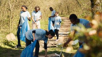 Diverse group of activists gathering to clean up a forest area, using litter cleanup tools like garbage bags and a long claw to grab the trash. People picking rubbish and recycling. Camera A. photo