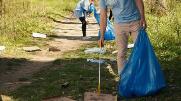 Young volunteer doing litter cleanup with a long claw to grab trash and store it in a garbage bag, dealing with pollution an illegal dumping problem. Activist protecting natural ecosystem. Camera A. photo