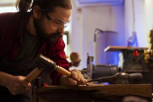 Man shaping raw timber using chisel and hammer in carpentry shop, wearing safety glasses to avoid injury. Woodworking expert making wood sculptures using protective gear to prevent workplace accidents photo