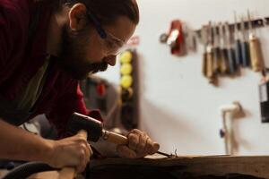 Sculptor shaping raw timber using chisel and hammer in carpentry shop, creating wood art, wearing safety glasses. Artist making wood sculptures, engraving lumber with tools, using protective gear photo