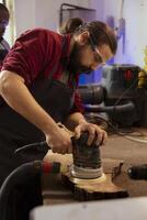 Skilled carpenter in joinery using orbital sander on lumber block with precision. Woodworker in carpentry shop smoothing rough wooden surfaces with professional angle grinder equipment photo