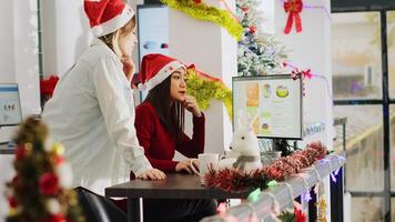 Employee wearing Santa hat working in Christmas ornate office requesting help from coworker. Staff member looking over business revenue numbers with colleague in xmas adorn workplace, handheld camera photo