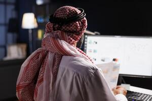 Focused Arab businessman diligently works at his desk, conducting online tasks and examining paperwork. Over-the-shoulder shot of a Muslim guy holding papers and using his modern desktop computer. photo