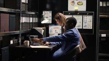 Diverse detectives in well-organized office reviewing evidence, surrounded by shelves filled with files. Two law enforcement officers with laptop, diligently investigate criminal profiles and records. photo