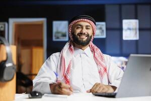 Portrait of a male Arab businessman looking at the camera, working at a desk with his digital laptop close by. Front-view shot of smiling Muslim guy writing on a notepad using a pen. photo
