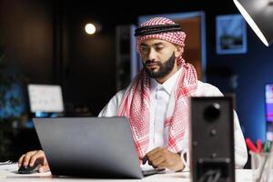 Arab businessman with confidence typing on a laptop at a contemporary desk. Male Muslim person using his personal computer for email and browsing in a serene and professional workplace environment. photo