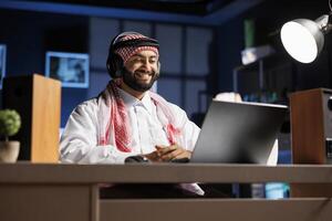 Handsome man in traditional Islamic attire, sitting at desk with laptop, smiles as he does research online. Arab guy with wireless headphones engrossed in conferencing and email communication. photo