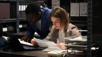 Multiethnic law enforcement agents conducting an investigation analyzing classified documents and evidence. Caucasian law officer uses laptop and discusses with policeman working towards justice. photo
