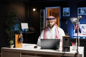 At modern workstation, confident Arab businessman types on his laptop. Muslim guy is browsing, conversing through email, and using WiFi connectivity in a calm and professional business environment. photo