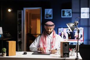 Middle Eastern guy dressed in Arabic attire sits at a table with his digital laptop, prepared for work. Committed Arab man plans to set up his personal computer to browse the internet. photo