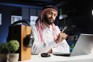 Close-up of an Arabian businessman dressed traditionally working intently at his contemporary desk, utilizing wireless technology and internet connection for research and conferencing. photo