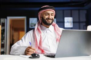 Smiling Arab guy works diligently at office desk, engages in digital tasks and writes on his notepad with laptop nearby. He efficiently conducts online research, embodying dedication and productivity. photo