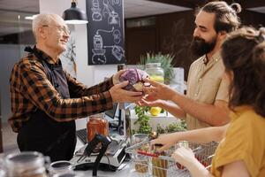 Older man at checkout counter in zero waste shop selling healthy vegetables in jar to vegan customers with green living lifestyle. Hipster couple buying groceries in local neighborhood store photo