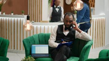 Bellboy talking on phone and taking notes, sitting in lobby at five star hotel. Concierge calling guests to ensure easy check in with all personal information, assisting visitors in arranging room. photo