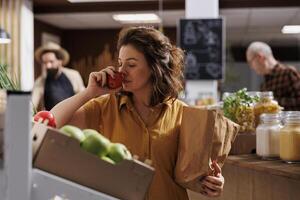 Woman smelling organic farm grown apples in busy zero waste store with low carbon emissions. Client testing to see if local grocery supermarket fruits are fresh and pesticides free photo