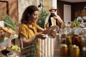 Zero waste shop storekeeper at checkout counter bagging organic bio vegetables grown locally, while customer in blurry foreground looks for pesticides free healthy groceries photo