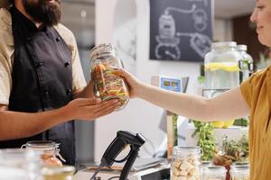 Vendor at checkout counter in zero waste shop selling nutritious produce in jar to customer with green living lifestyle. Client buys ethical locally grown food in local neighborhood store, close up photo