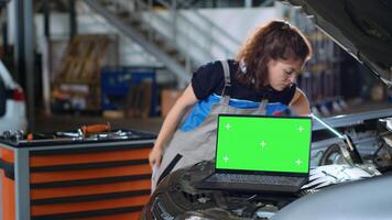 Chroma key laptop in garage workplace sitting on opened up car. Mockup device in auto repair shop next to mechanic cleaning vehicle inside engine compartment after fixing it photo
