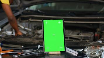 Close up shot of tablet placed on working bench in busy garage next to professional tools while engineer fixes vehicle. Mockup device in repair shop with precise expert working in blurry background photo