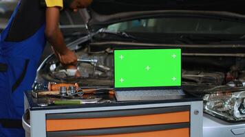 Close up shot of laptop placed on working bench in busy garage next to professional tools while serviceman fixes vehicle. Mockup device in auto repair shop with mechanic working in blurry background photo