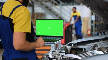 Licensed repairman using green screen laptop in car service to order new parts for busted vehicle. Close up shot of professional using chroma key device to look on internet for replacing components photo