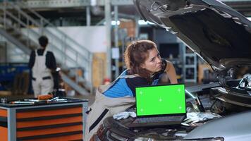 Chroma key laptop in garage facility sitting on malfunctioning car. Green screen device in auto repair shop next to mechanic cleaning vehicle inside engine compartment after fixing it photo