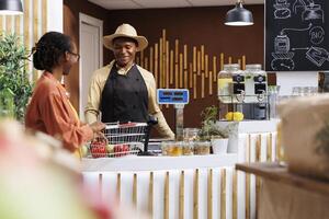 Friendly black male cashier with warm smile assists female client with her purchase at local market checkout counter. The image showcases excellent customer service and diverse shopping experience. photo